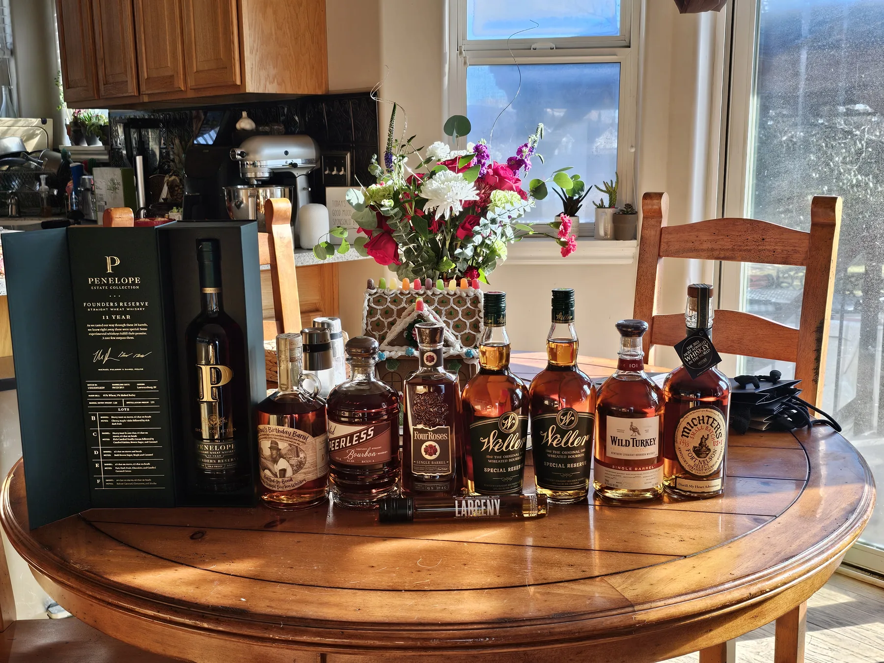A row of whiskey bottles on a round wooden dining table with flowers behind them