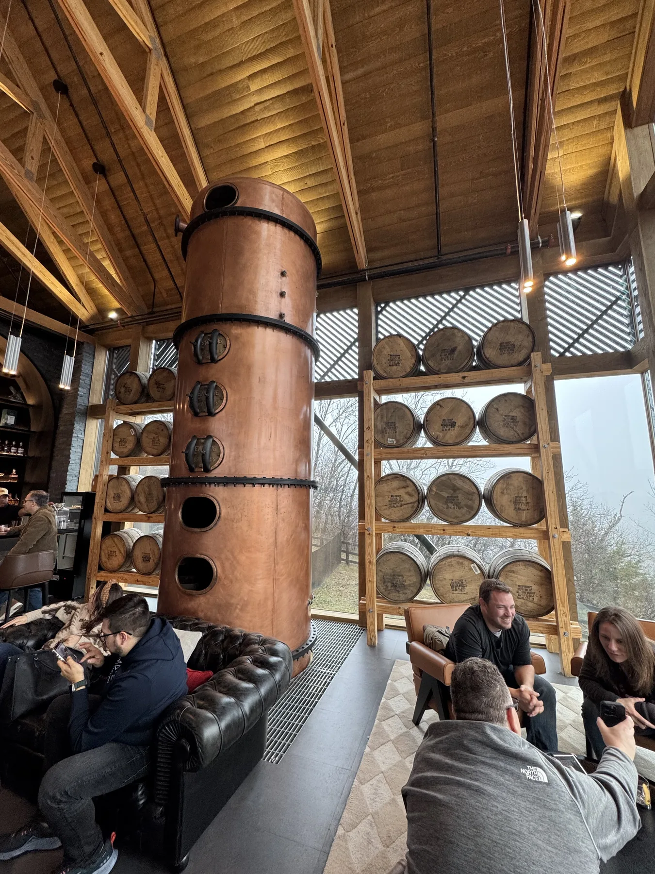 Visitors seated in a distillery lounge beside a tall copper still and racks of barrels